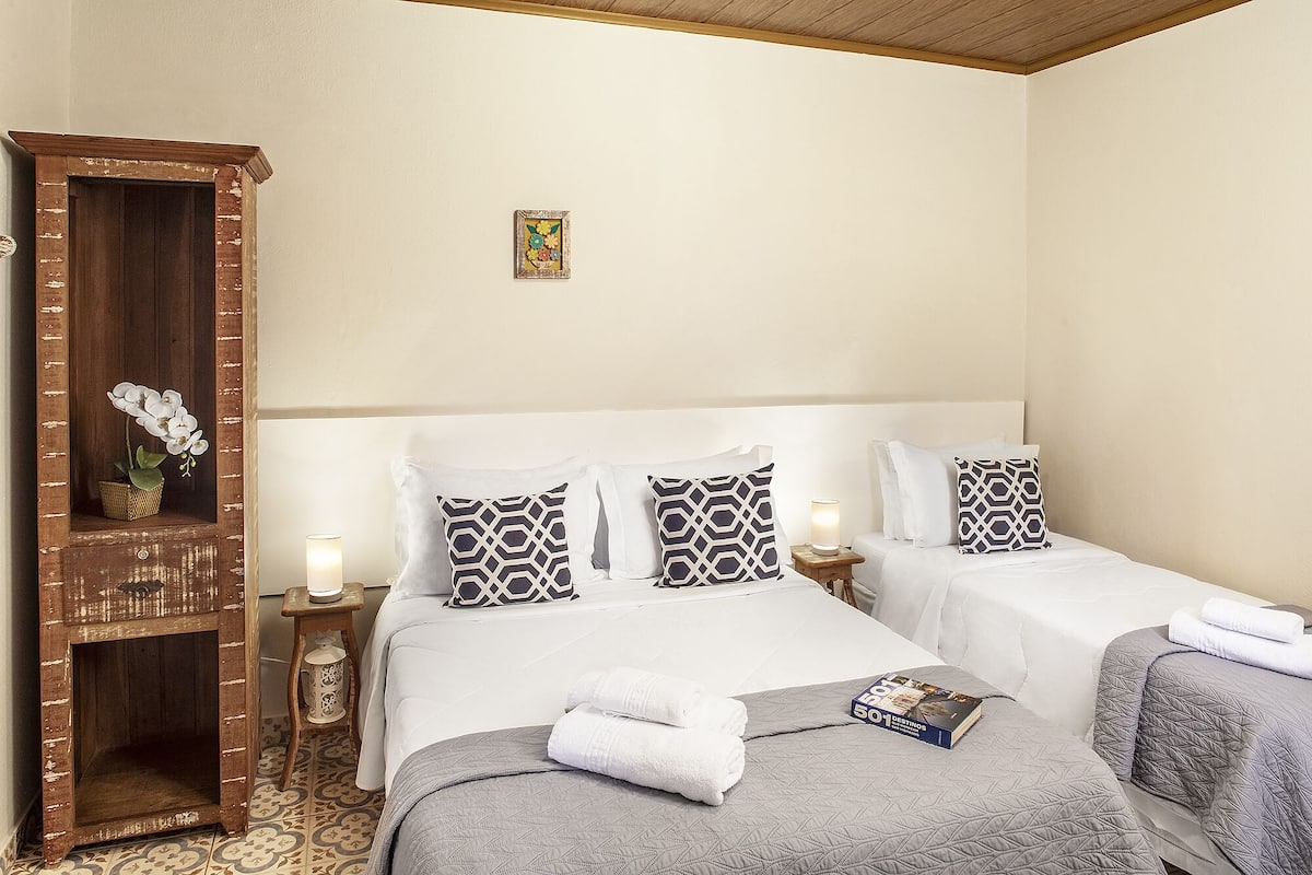 Quarto de hotel com duas camas, um armário de madeira e uma mesinha com um candeeiro.