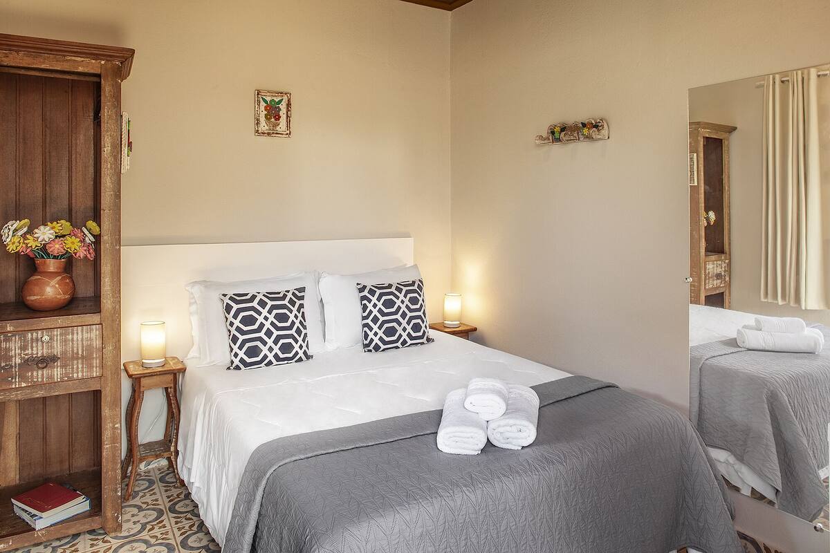 Uma cama bem arrumada com um cobertor cinza e duas almofadas, uma mesa de cabeceira com abajur, e um armário de madeira com um vaso de flores.
