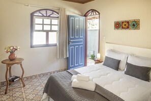 A bedroom with a bed, a window, a door, a small table with a vase, and a wall decoration.