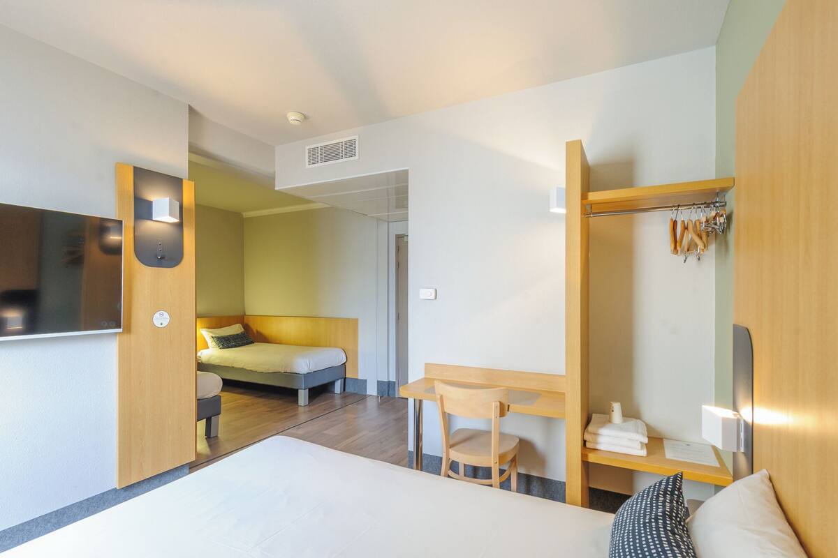 Ein modernes Hotelzimmer mit Bett, Schreibtisch, Stuhl, Fernseher, Kleiderhaken und einer Badezimmertür.