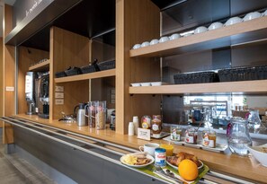 Una barra de desayuno con máquinas de café, cestas y una variedad de alimentos para el desayuno, incluyendo huevos, tostadas y zumo de naranja.