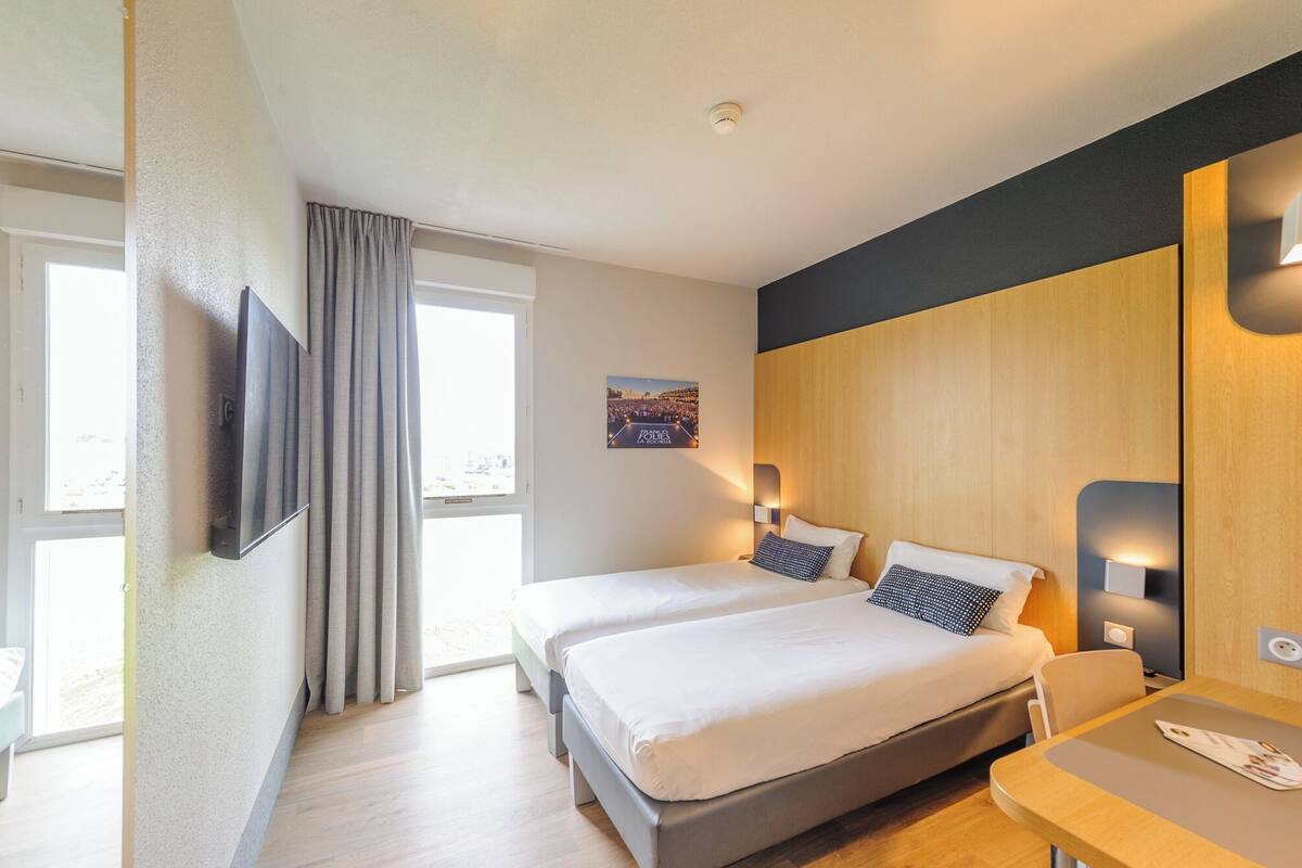 Ein Hotelzimmer mit zwei Betten, einem Schreibtisch, einem Stuhl, einem Fernseher und einem Fenster mit Vorhängen.