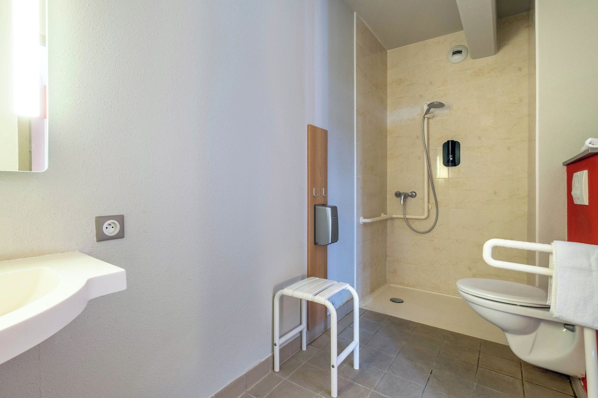 A bathroom with a white sink, a small white stool, a shower with a handheld showerhead, and a toilet.