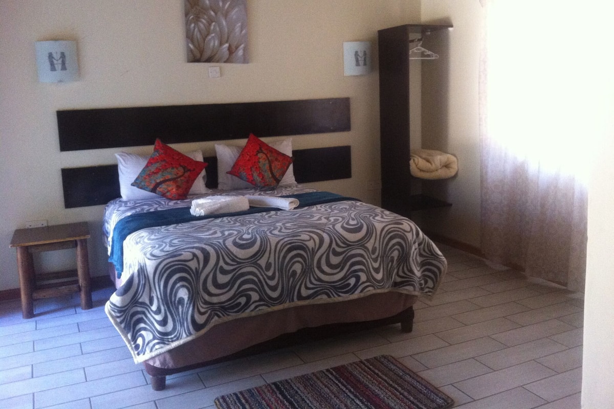 A neatly made bed with a patterned bedspread and two red pillows, a small bedside table, and a wall-mounted artwork.
