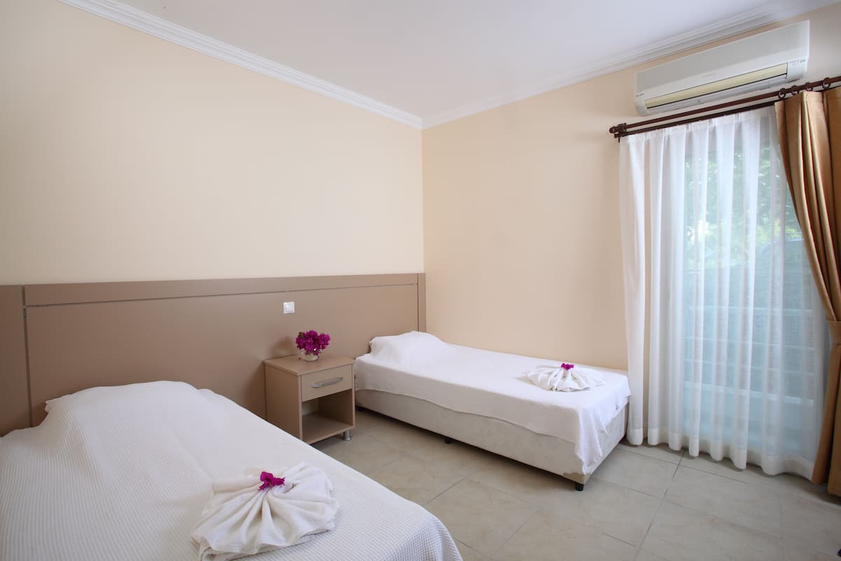A hotel room with two single beds, a small bedside table with a vase of flowers, a window with sheer curtains, and an air conditioning unit above the window.