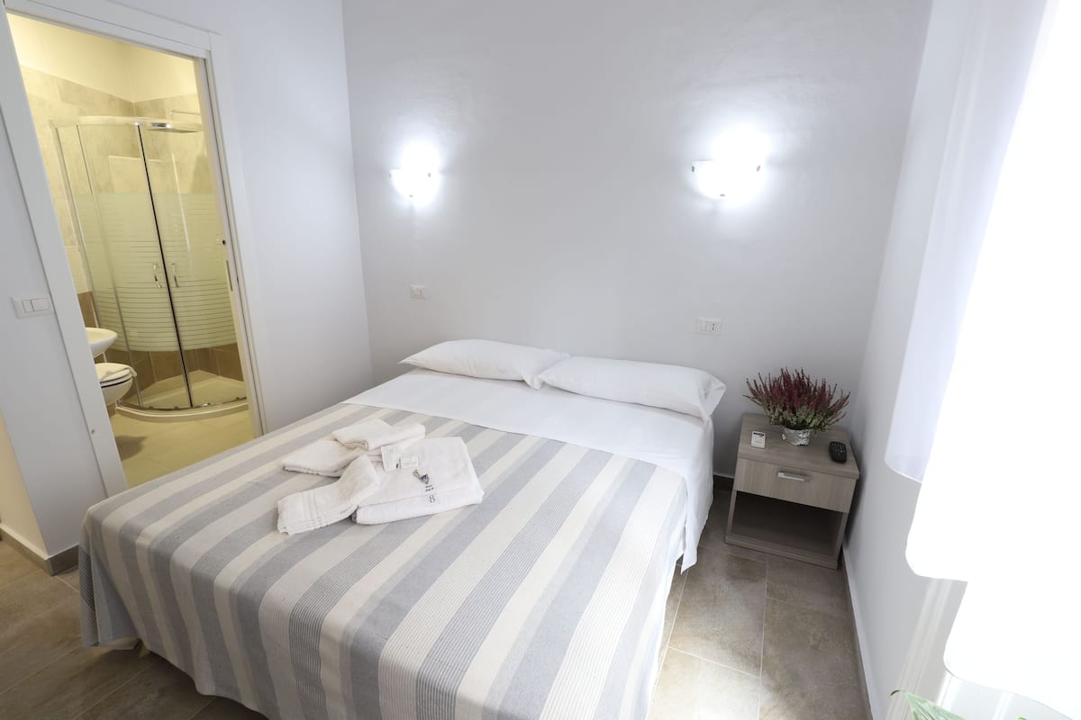 A hotel room with a bed, a nightstand, a small table with a plant, a shower area, and a toilet.