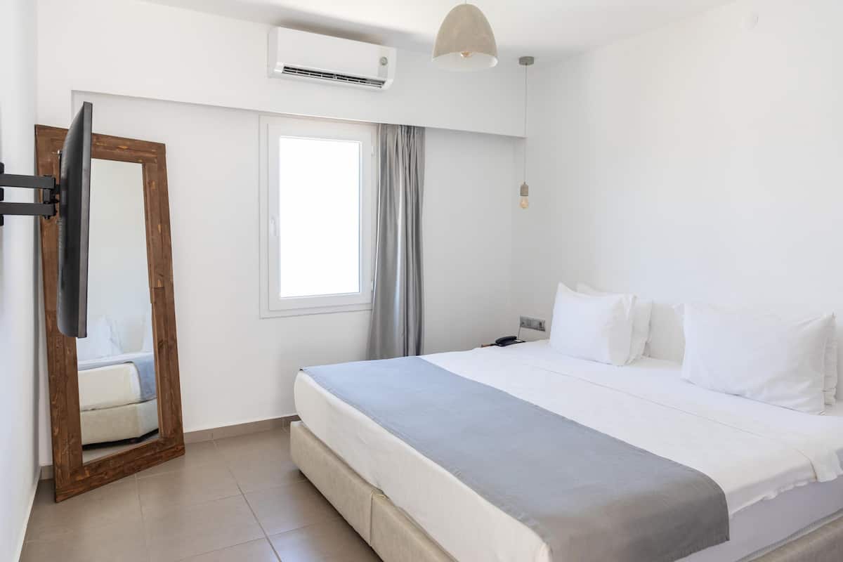 A hotel room with a bed, a mirror, a window with curtains, an air conditioner, and a pendant light.