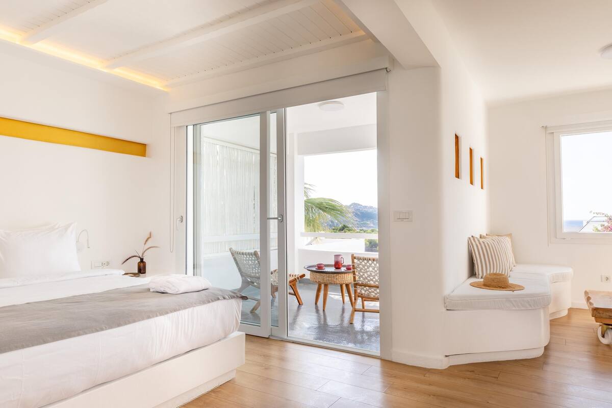 A modern bedroom with a bed, a balcony with a table and chairs, and a view of the outdoors.