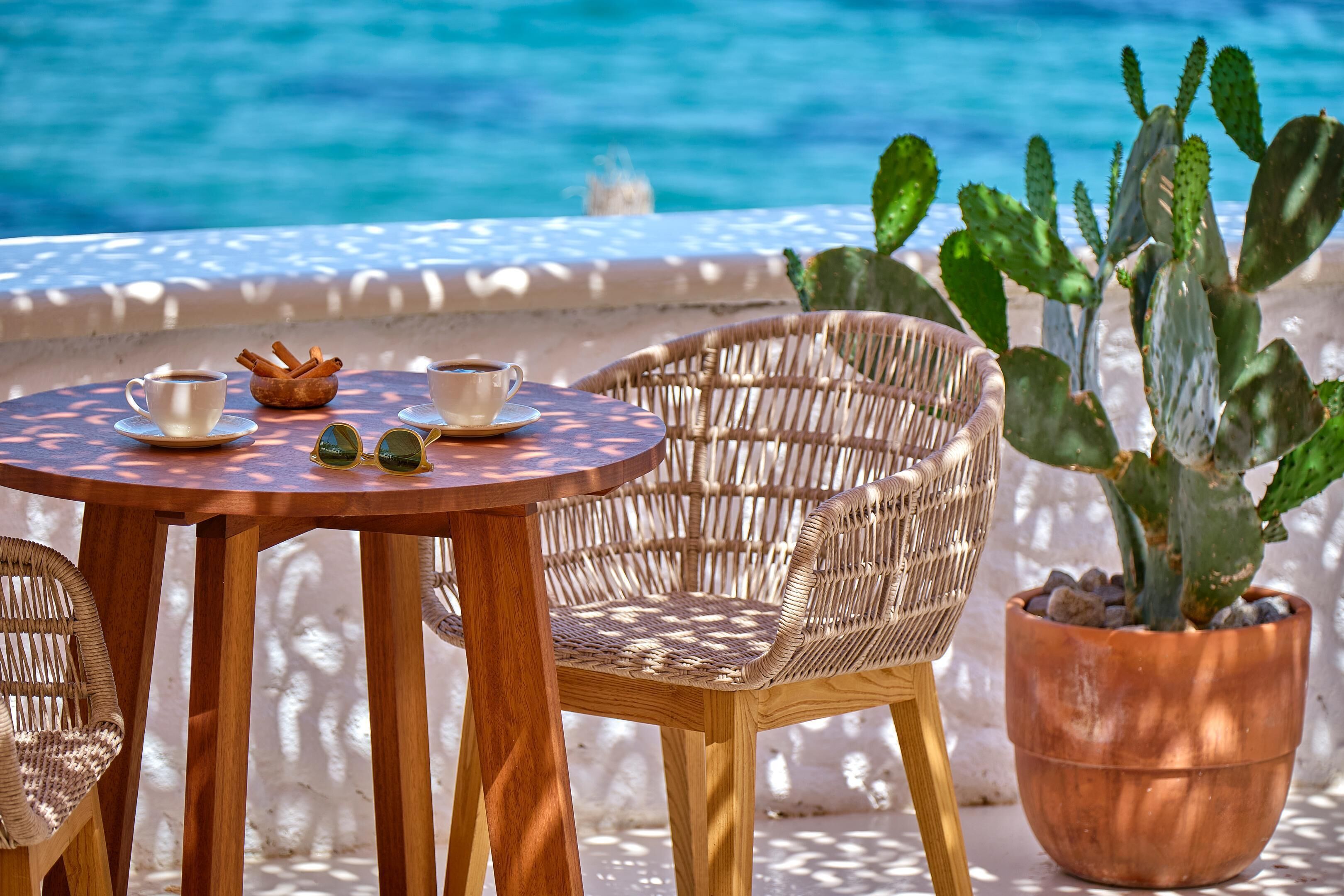 A round wooden table with two chairs, a potted cactus, and two cups of coffee on a terrace with a view of the sea.
