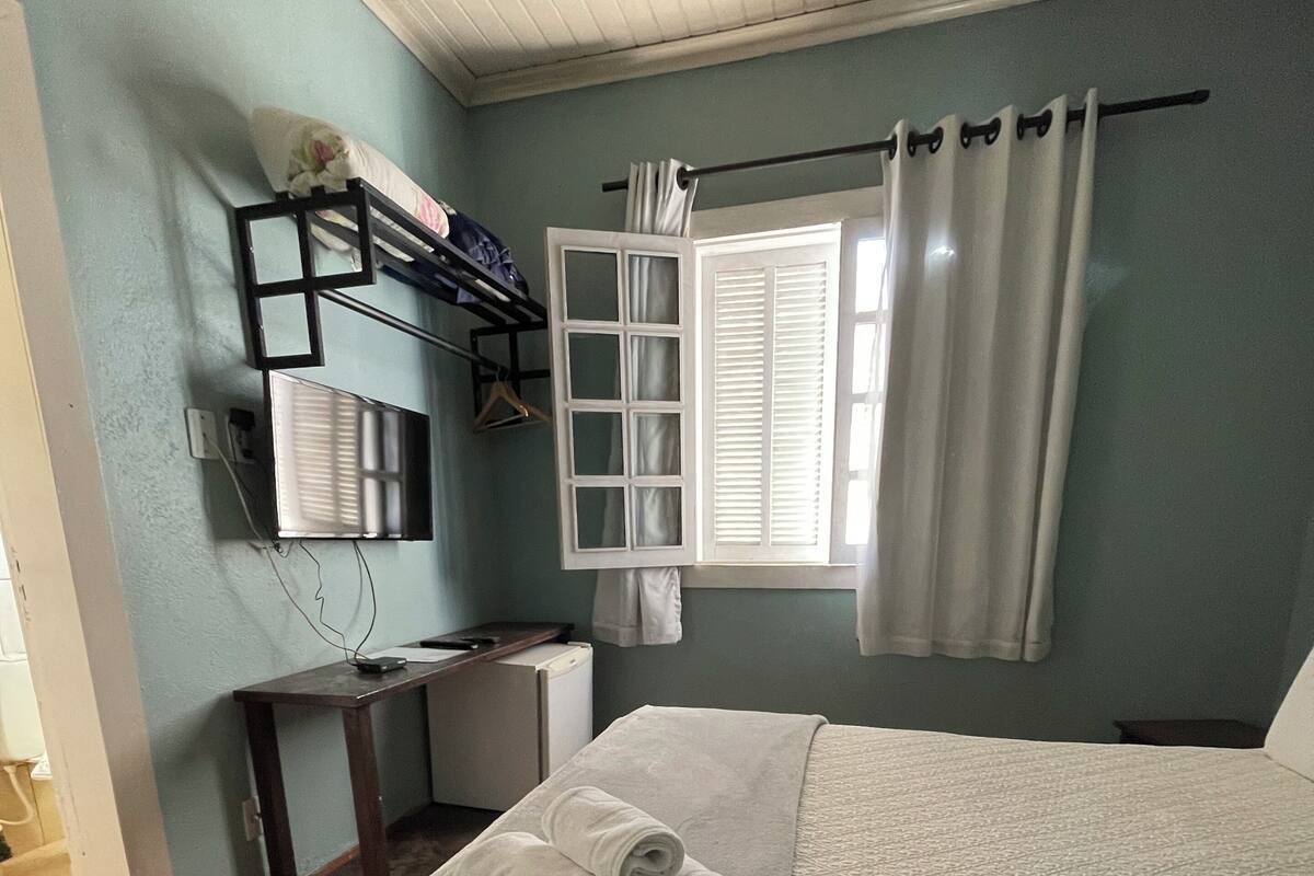 Un lit bien fait, recouvert d’une couette blanche et agrémenté de deux serviettes enroulées, une petite table de chevet avec une lampe, un téléviseur fixé au mur et une fenêtre avec des rideaux blancs.