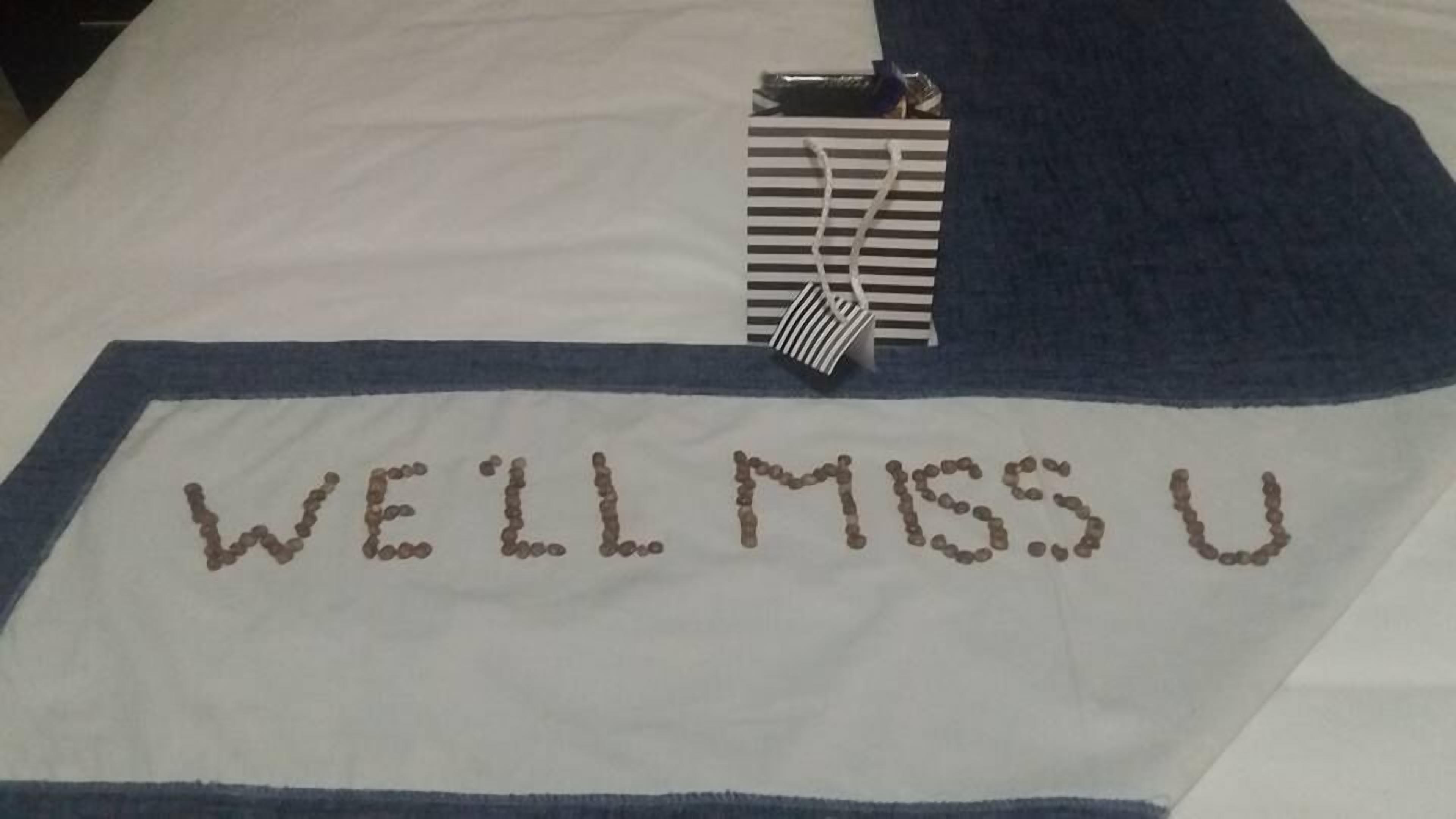 A bed with a blue and white blanket. The blanket has the text "WE'LL MISS U" embroidered in brown.