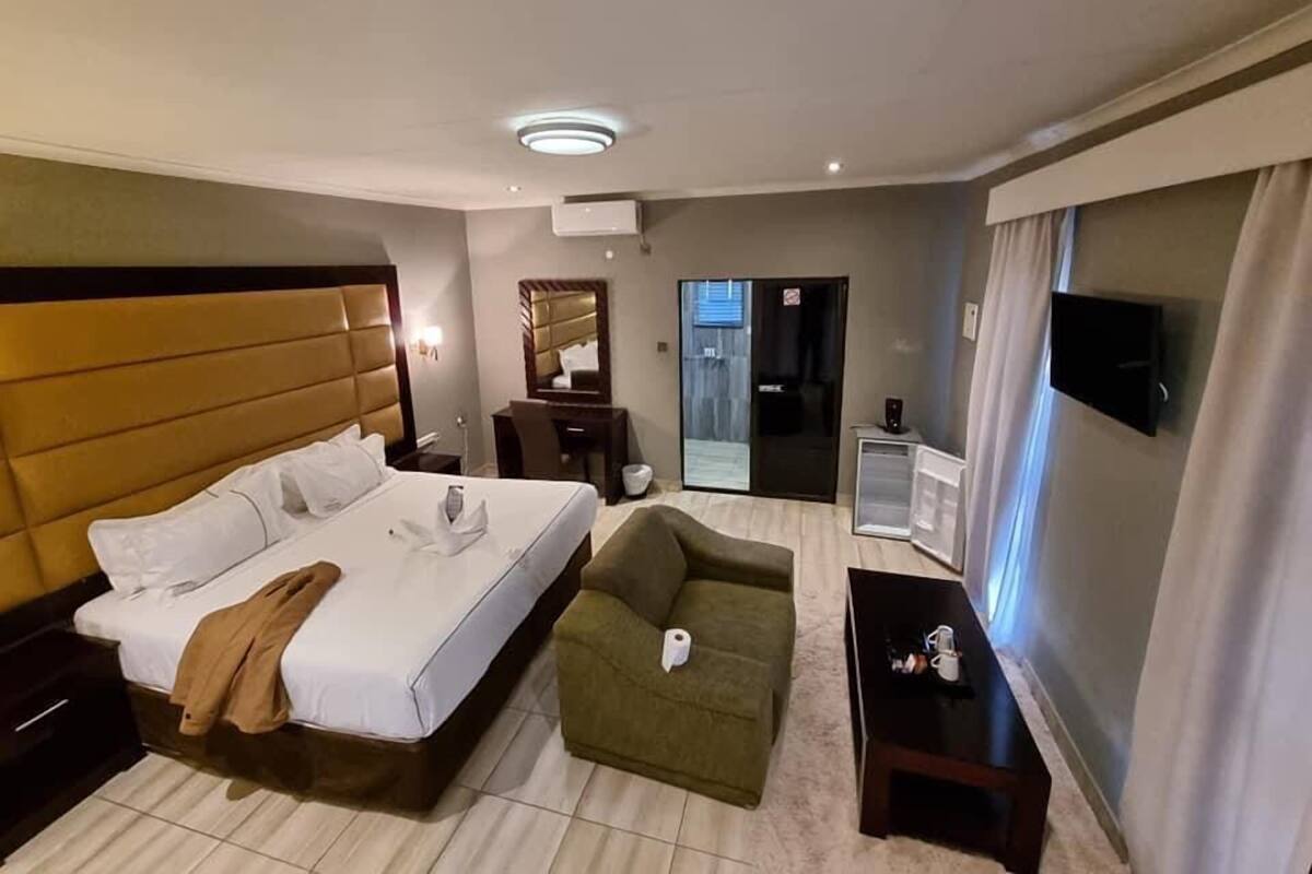 Habitación de hotel con una cama grande, un sillón verde, una mesita, un televisor y una puerta que da a un balcón.