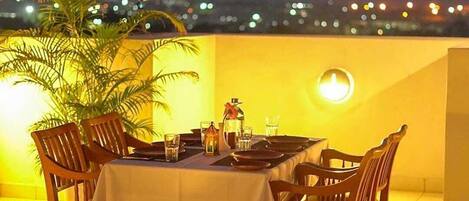 Un área de comedor al aire libre con una mesa dispuesta para cuatro personas, rodeada de sillas de madera, bajo una iluminación amarilla y una vista panorámica de la ciudad de fondo.