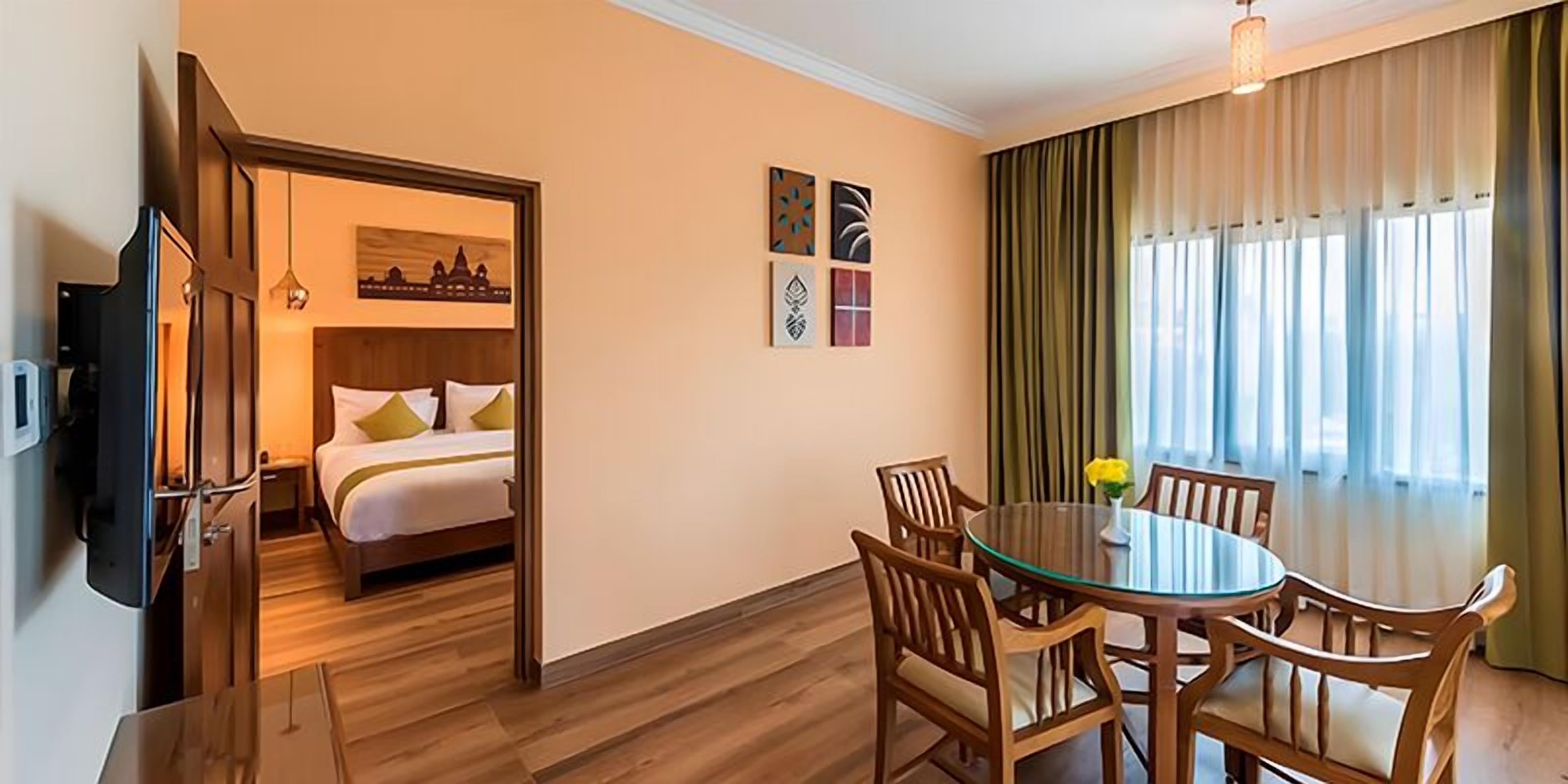 A hotel room with a bed, a dining table with chairs, a TV, and artwork on the wall.