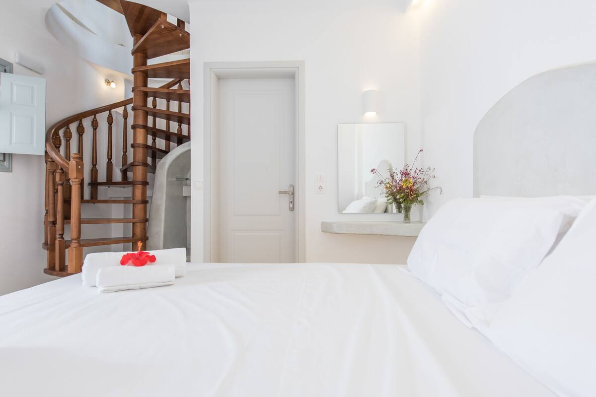A hotel room with a wooden spiral staircase, a white door, a bedside table with a vase of flowers, and a neatly made bed.