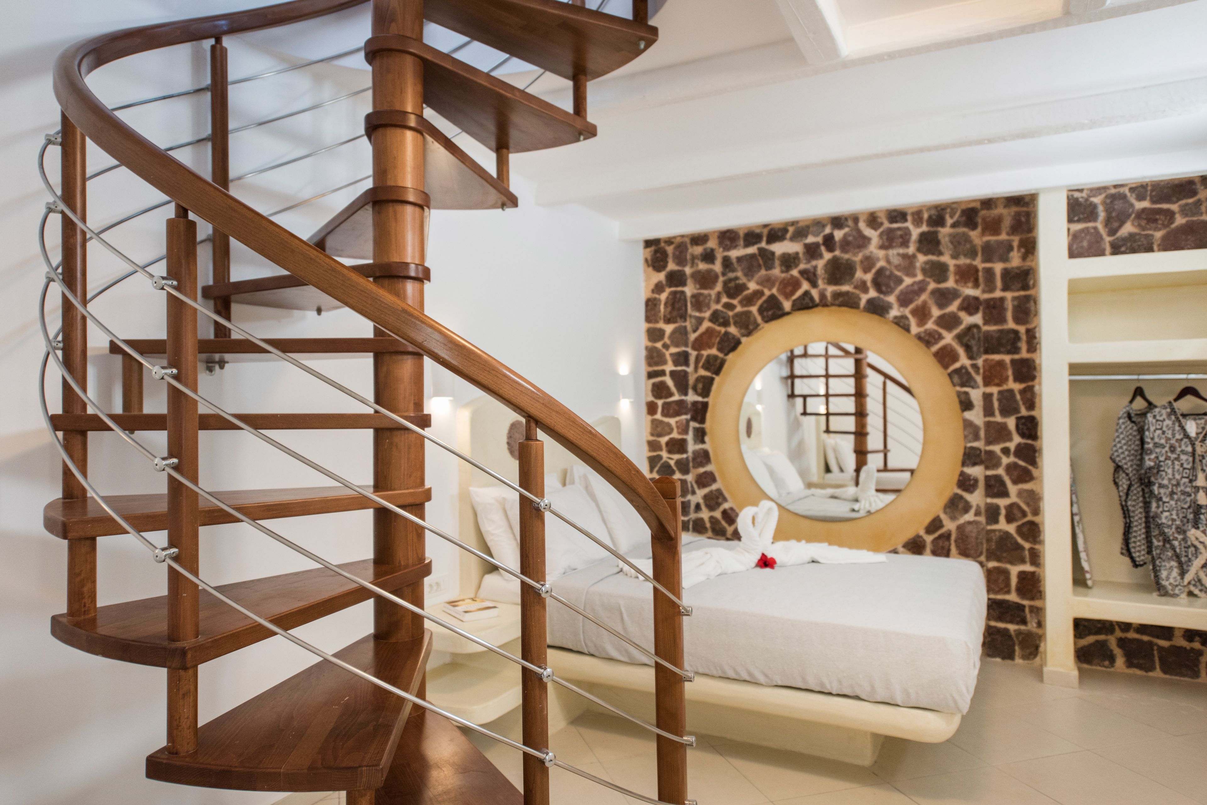 A spiral staircase with wooden railings and metal handrails, leading to a bedroom with a stone accent wall and a round mirror above the bed.