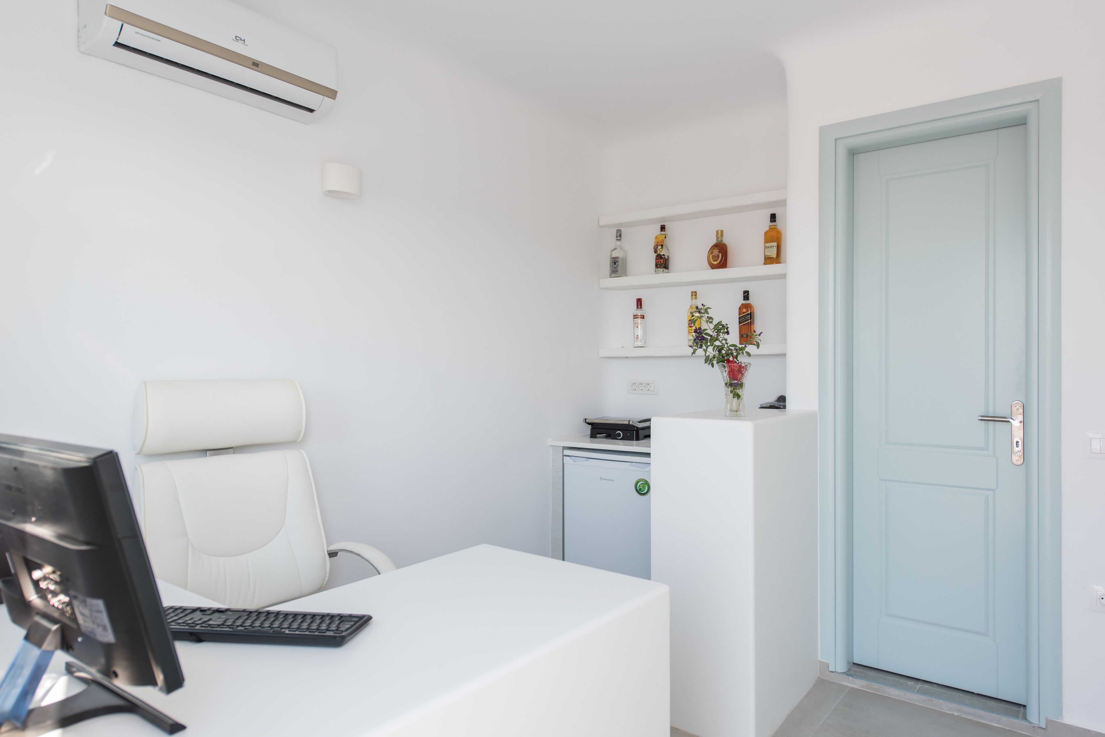 Una oficina moderna con un escritorio blanco, computadora y silla. Hay un refrigerador pequeño y estantes con botellas. Se ve una puerta azul claro.