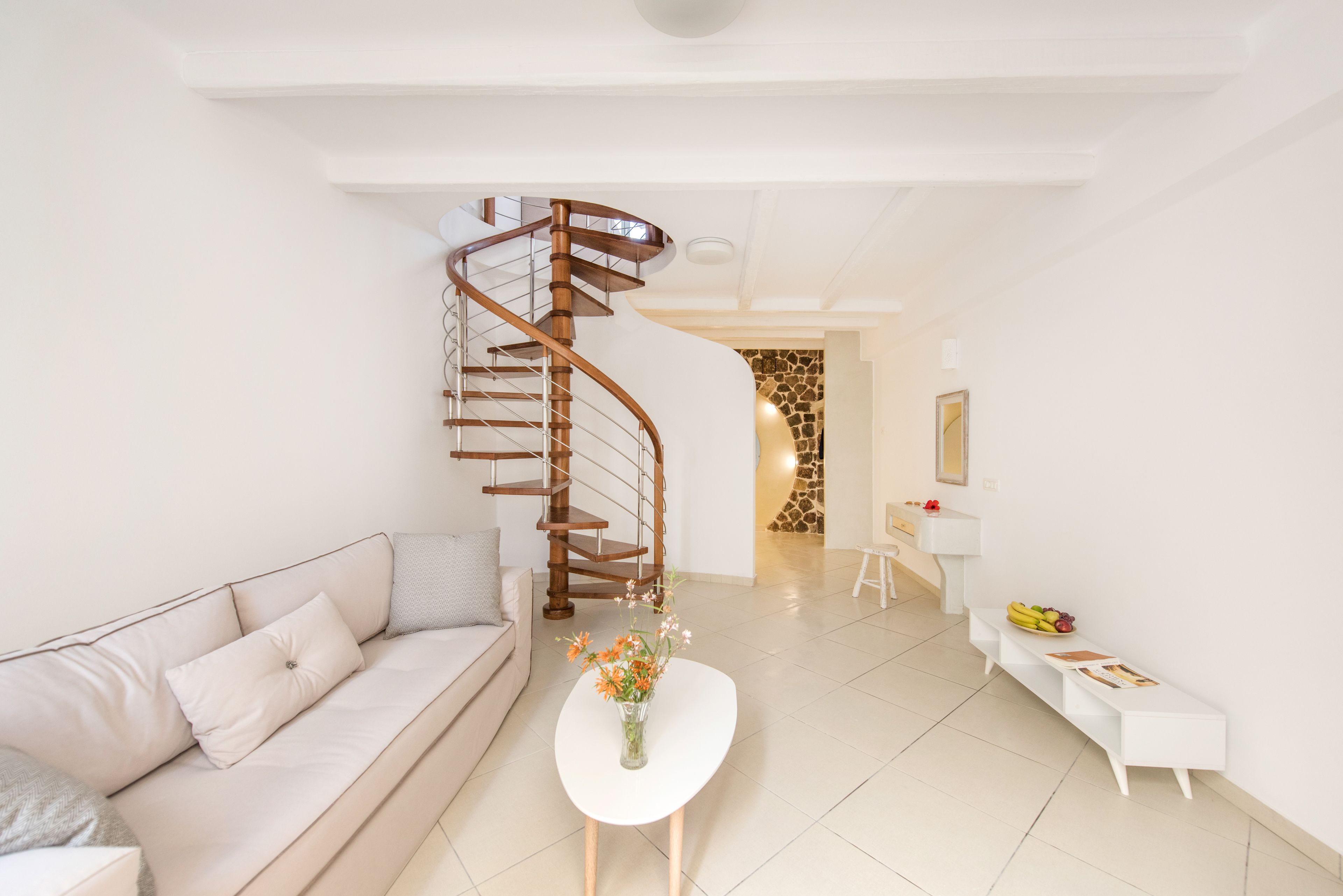 A modern living room with a spiral staircase, a white sofa, a coffee table with flowers, and a white console table with fruits and magazines.