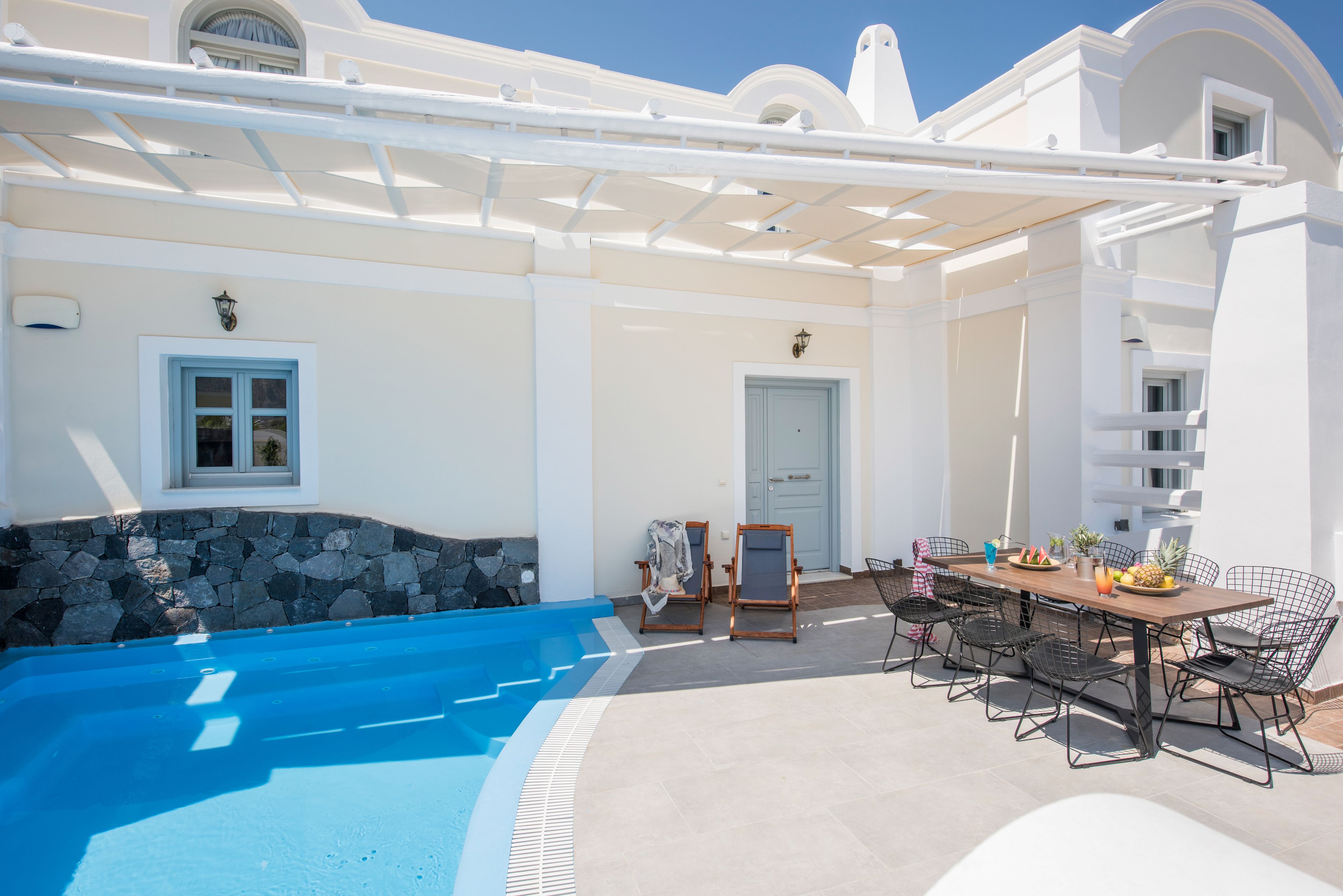 Un'area piscina con muro in pietra, una zona relax coperta con tavolo e sedie, e una porta che dà su un edificio.