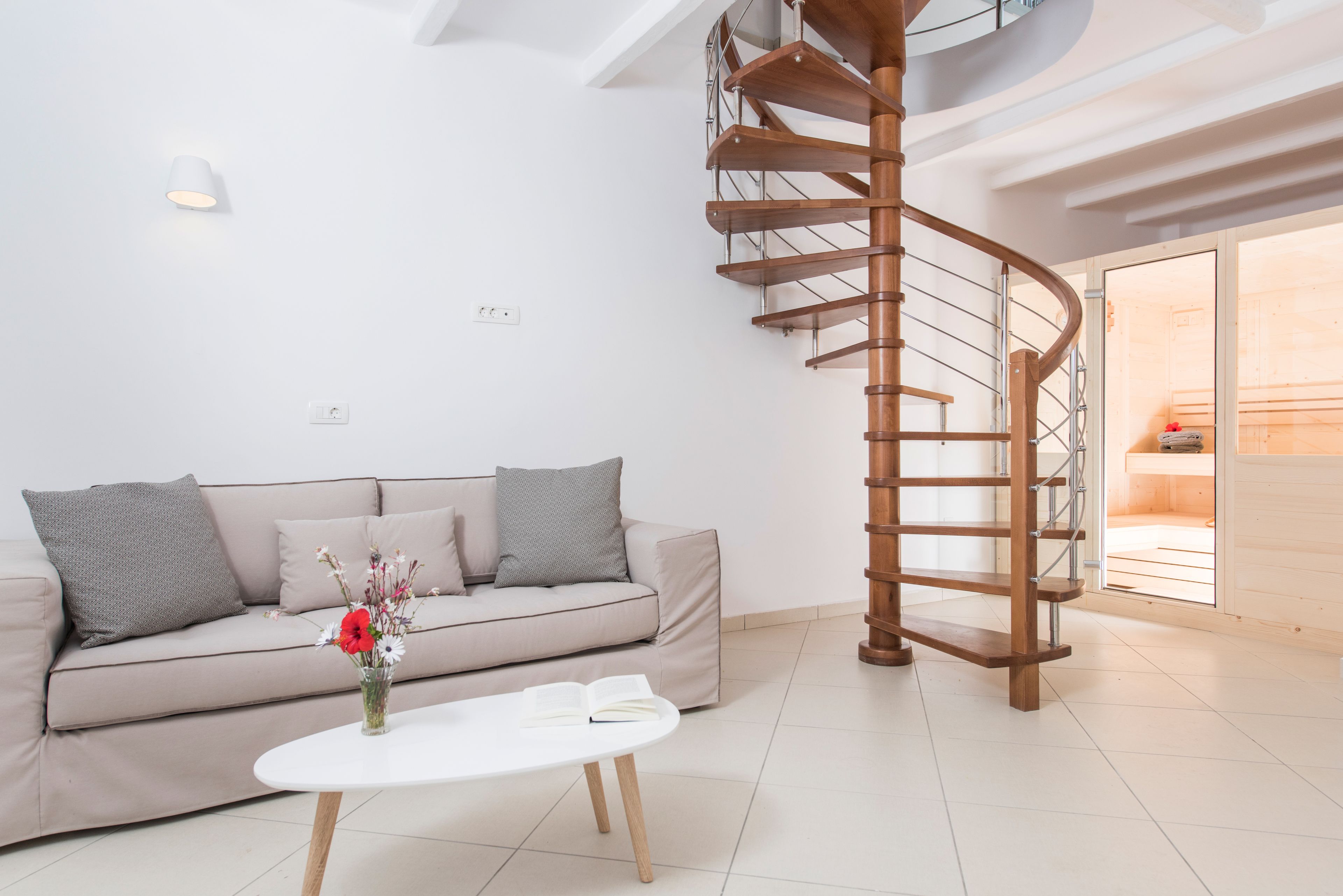 A modern living room with a spiral staircase, a sofa with cushions, a white coffee table with a book and flowers, and a light fixture on the wall.