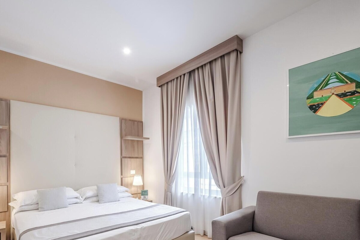 A hotel room with a bed, a chair, a painting, and a window with curtains.