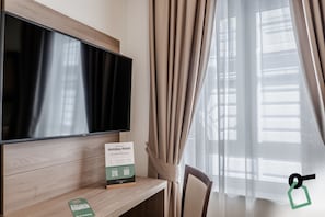 Una camera d'albergo con televisore a schermo piatto su un supporto in legno, un cartello sulla scrivania e tende trasparenti alla finestra.