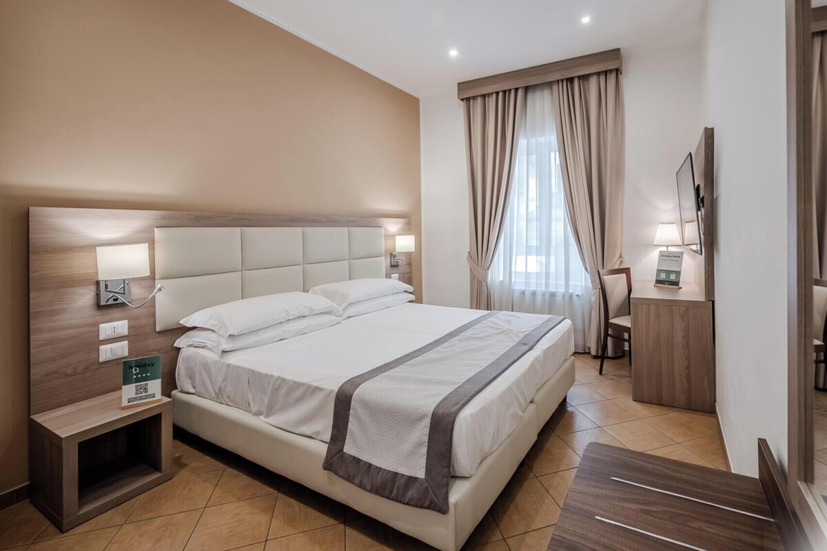 A hotel room with a large bed, bedside lamps, a nightstand, a desk with a lamp, a mirror, and a window with curtains.