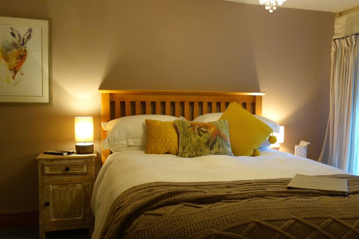 A neatly made bed with a wooden headboard, a bedside lamp, a nightstand, and a window with curtains.