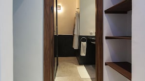Ein schmaler Flur führt zu einem Badezimmer, wo ein Handtuch an der Wand hängt und eine Regalvorrichtung sich rechts befindet.