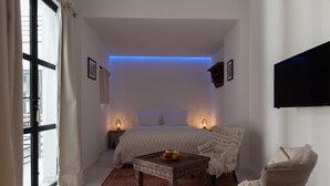 Ein Schlafzimmer mit einem Bett, einem kleinen Tisch mit Obst, einem Sessel und einem Fernseher an der Wand.