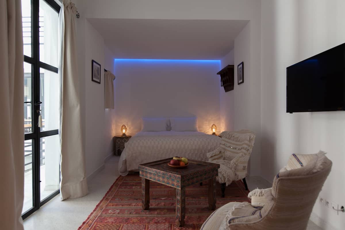 A bedroom with a bed, a small table with fruit, a chair, and a TV on the wall.