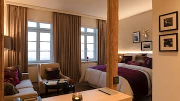 Quarto de hotel com uma cama, um sofá, uma cadeira, uma pequena mesa com uma vela e uma janela com cortinas.
