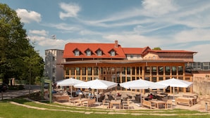A building with a red roof and large windows, outdoor seating with umbrellas, and a stone pathway leading to the entrance. - Hotel Brunnenhaus Schloss Landau (Bad Arolsen)