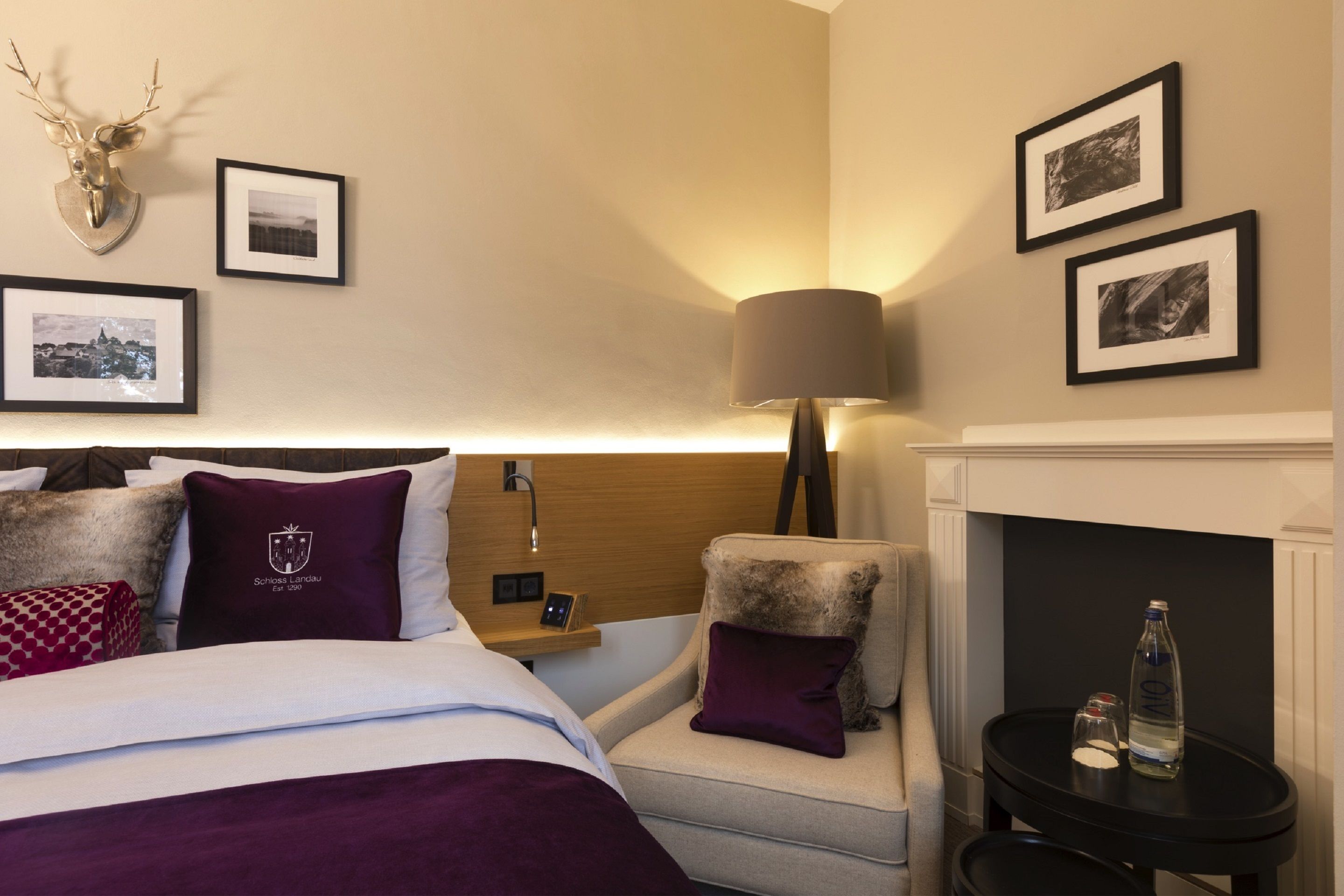 Una habitación de hotel con cama, un sillón pequeño, una mesita auxiliar, una lámpara, cuadros enmarcados en la pared y una chimenea.