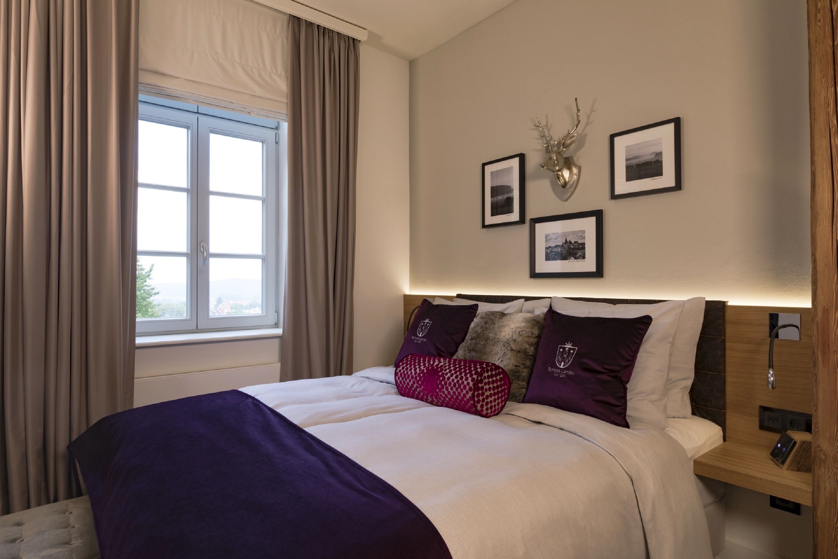 Una cama bien hecha con una colcha blanca y morada, una mesita de noche de madera con una lámpara y una ventana con cortinas.