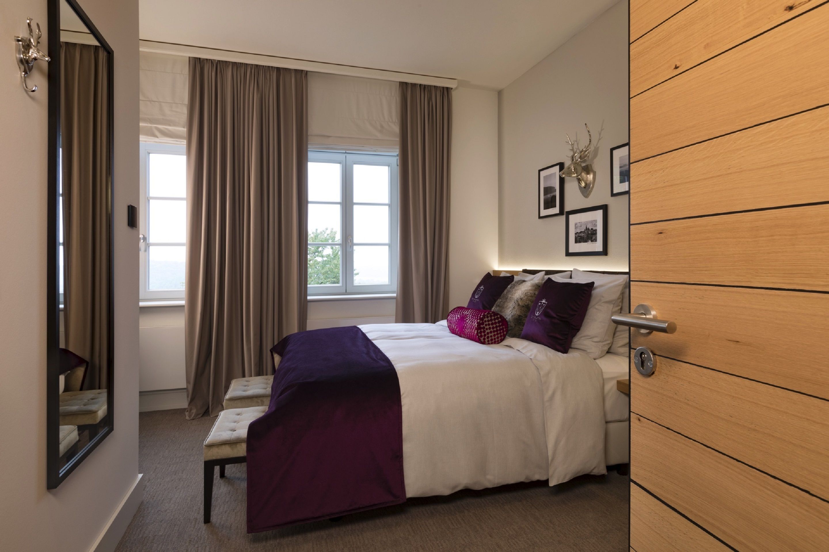 Un dormitorio con cama, un banco, un armario de madera, una ventana con cortinas y cuadros en la pared.