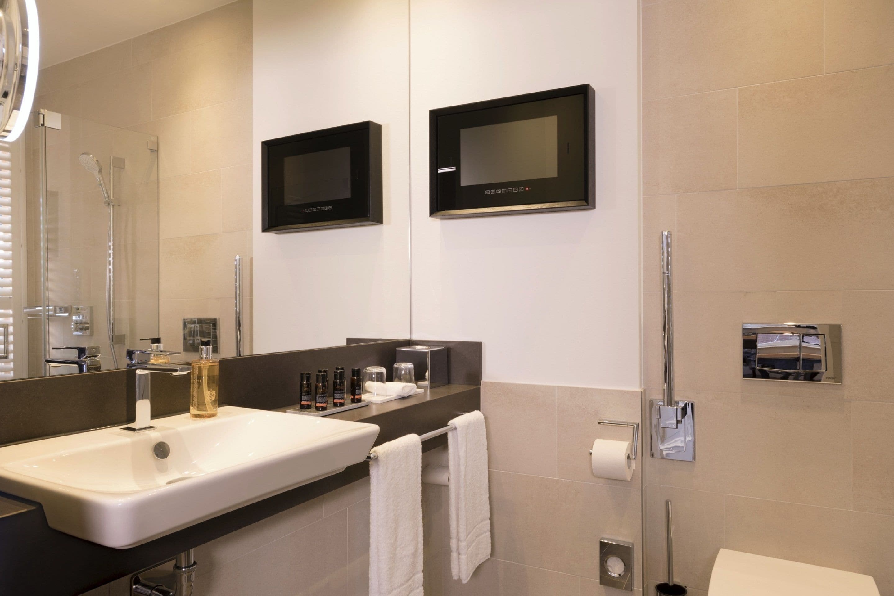 Un baño moderno con un lavamanos grande, dos televisores empotrados en la pared, un espejo y una zona de ducha.