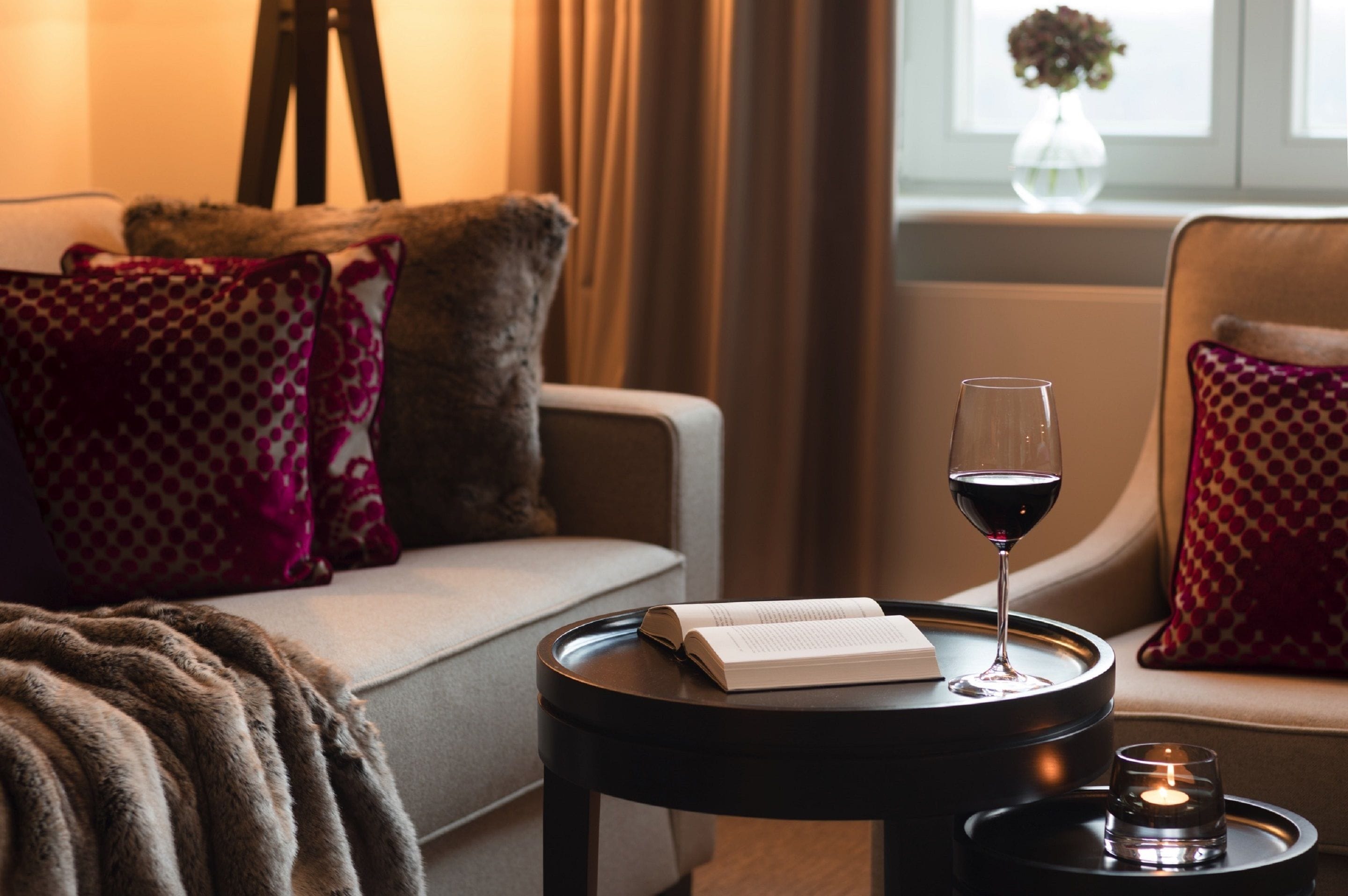 En mysig läshörna med en soffa, ett litet runt bord, en bok, ett glas rött vin och ett ljus.
