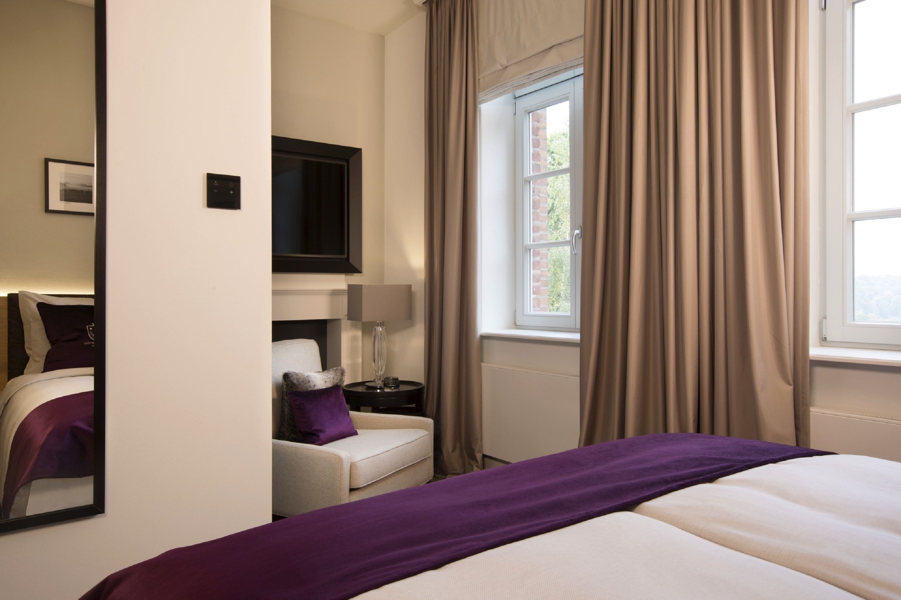 Una habitación de hotel con una cama, una silla, un televisor, un ventanal con cortinas y un panel de control en la pared.