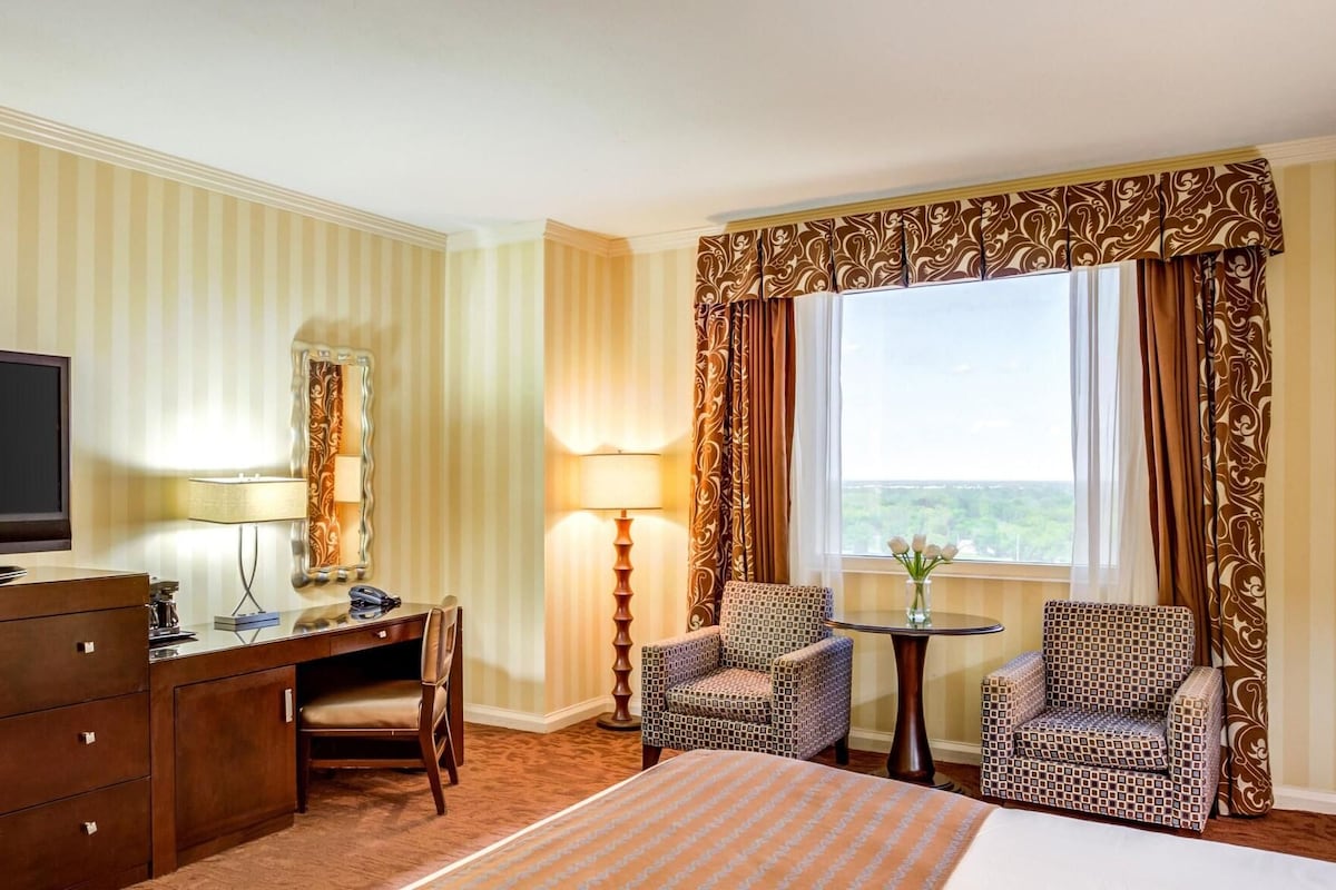 Une chambre d’hôtel comprenant un lit, un bureau avec une chaise, une télévision, une lampe et deux fauteuils à motifs.