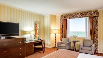 Quarto de hotel com cama, escrivaninha com televisão, duas poltronas estampadas, uma mesinha, abajur e janela com cortinas.