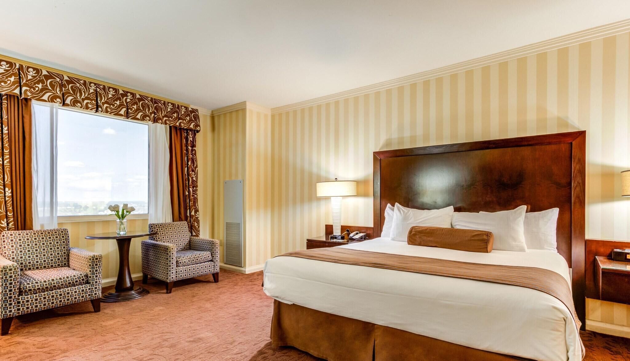 Une chambre d’hôtel avec un grand lit, deux fauteuils, une petite table et une fenêtre avec des rideaux.