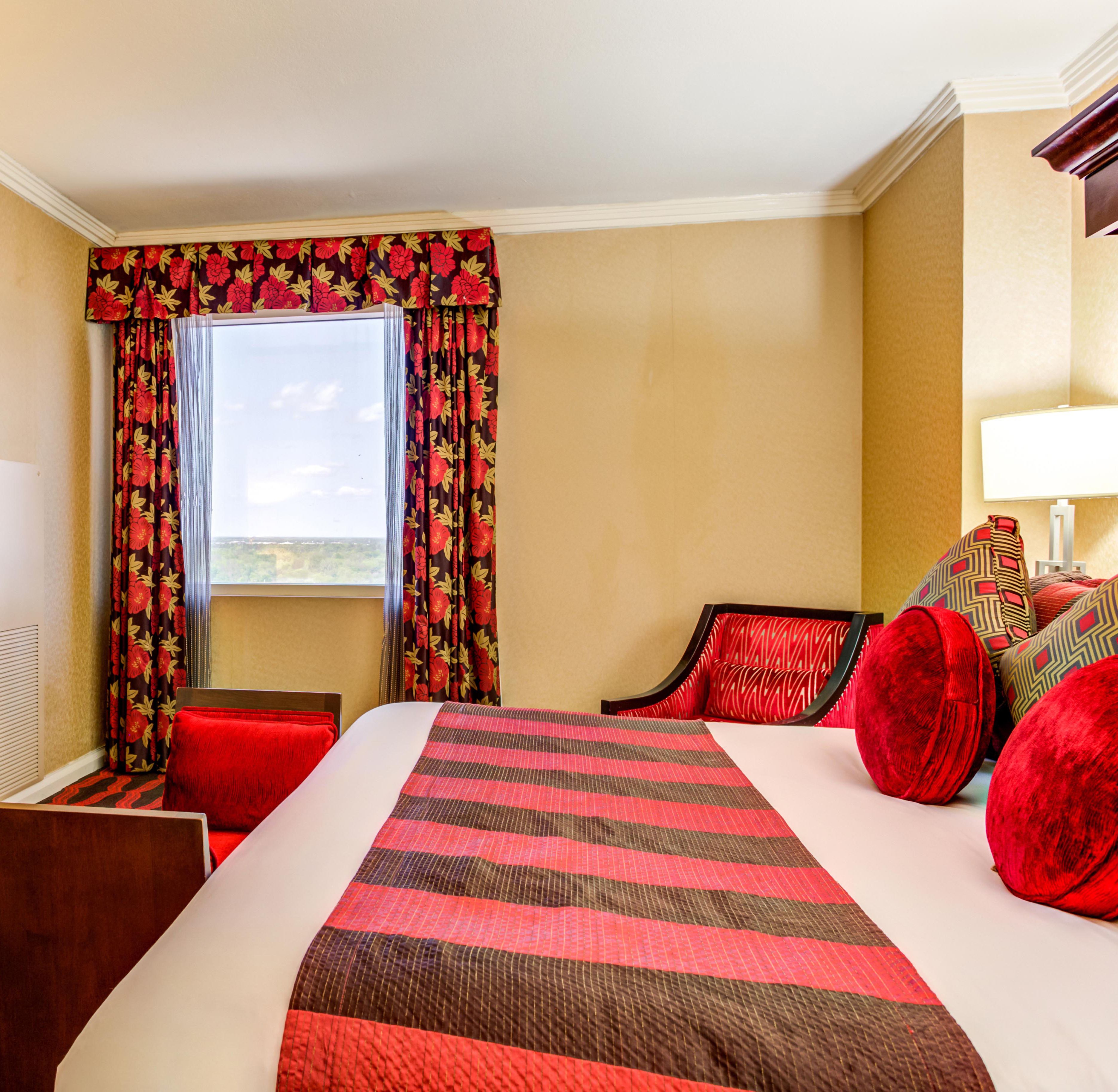 A hotel room with a large bed, red and black striped bedding, red floral curtains, and a view of the landscape through the window.