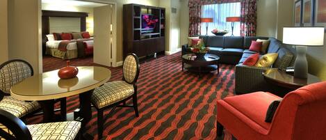 Een hotelkamer met een eethoek, een bank, een televisie en een groot raam met gordijnen.