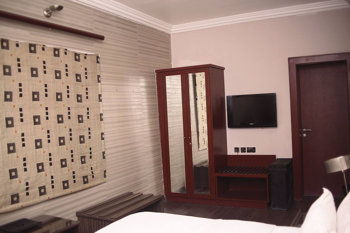 A hotel room with a bed, a wooden wardrobe, a television mounted on the wall, and a window with blinds.