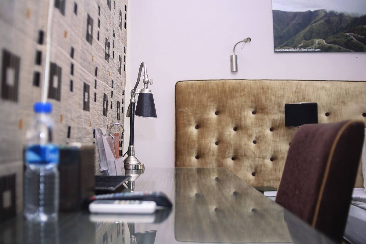 A hotel room with a tufted headboard, a glass table, a remote control, a water bottle, and a lamp.