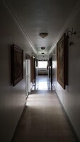 A long, narrow hallway with a tiled floor and white walls. There are framed pictures on the walls and a window at the end of the hallway.