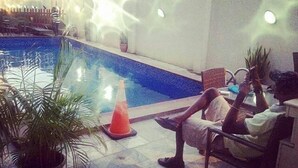 A pool area with a person sitting on a chair, a potted plant, and a traffic cone. - Capital Inn Plus (Ibadan)