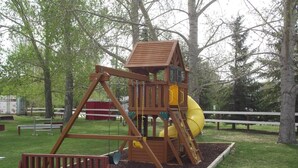 Un parque infantil de madera con un tobogán y una estructura para escalar, rodeado de árboles y una valla blanca.