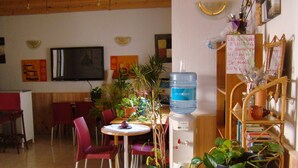 A dining area with a table, chairs, and a water dispenser. There are potted plants and a bookshelf with books and decorative items. - Hostal Platja (Cambrils)